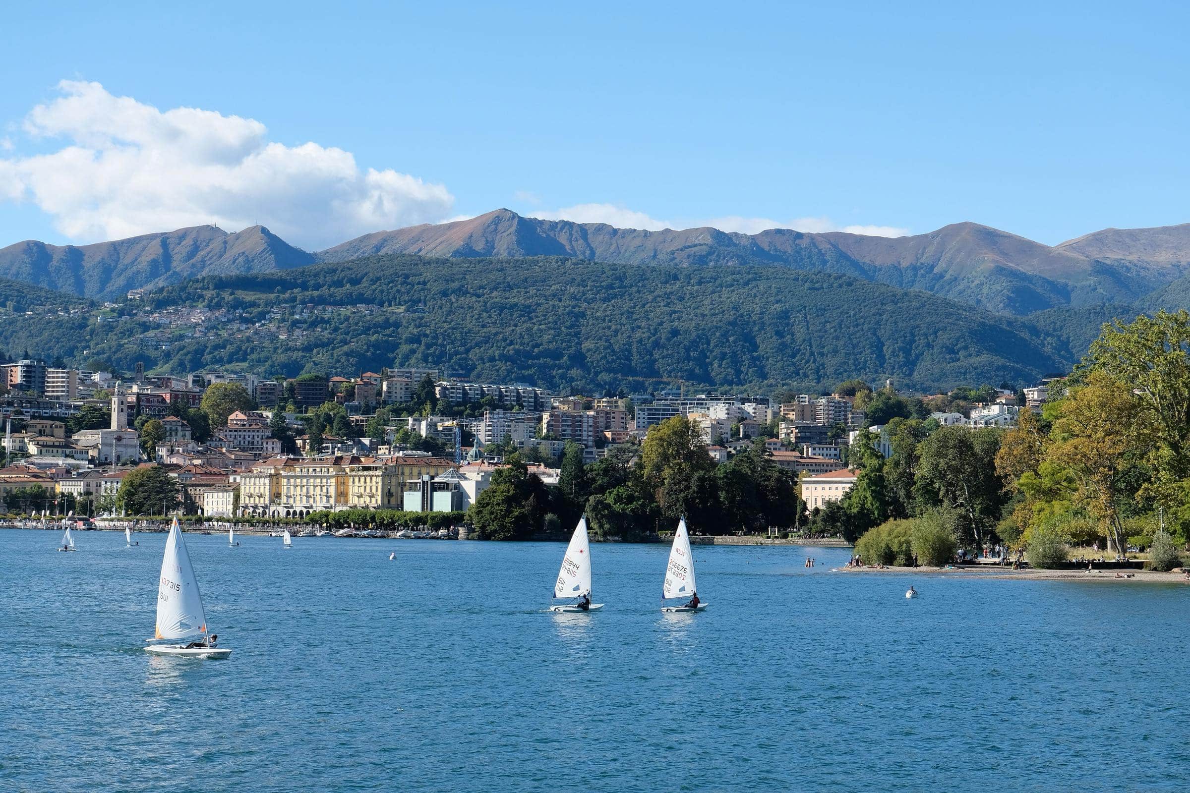Il golfo di Lugano in una giornata di sole: tre derive a vela sul lago, il lungolago e il centro città, le montagne sullo sfondo.